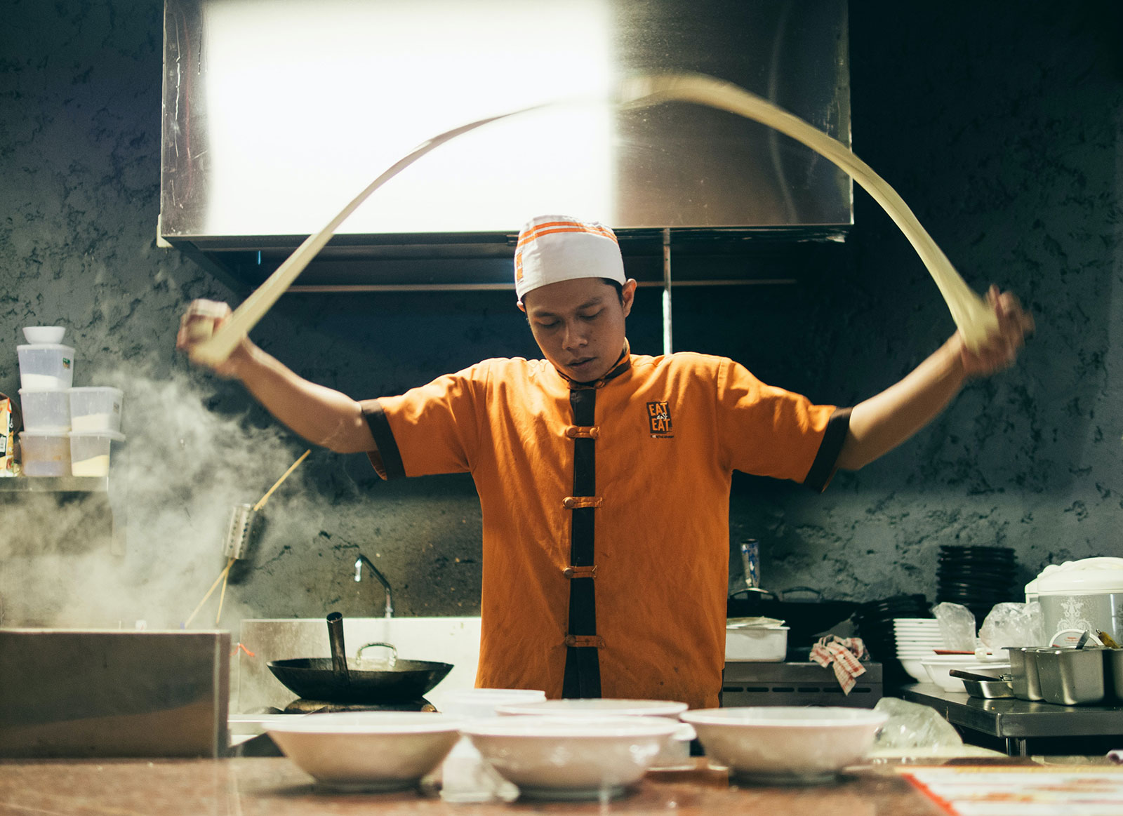 chef stretching noodle dough in air in kitchen surrounded by pots