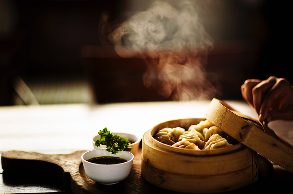 Dumplings in wood steamer basket and hand taking lid off allowing steam to come out