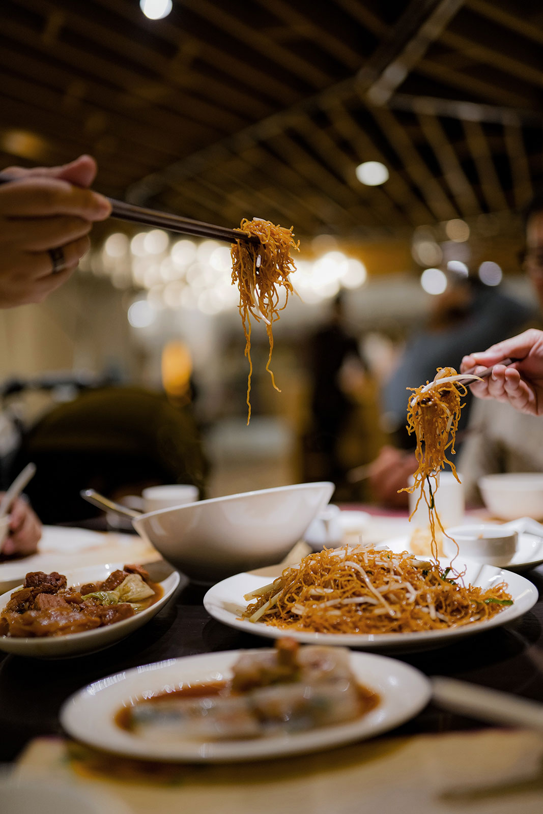 hands holding noodles with their chopsticks and plates of beef and noodle dishes on the table