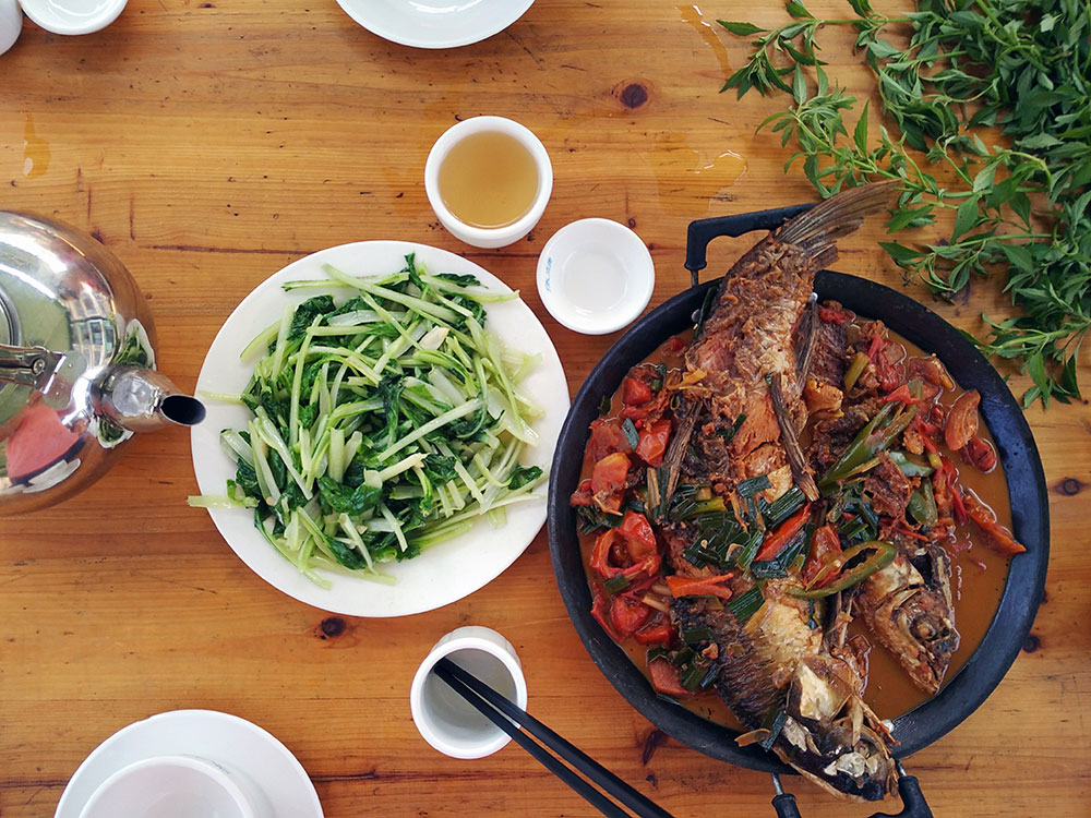 Two whole fish in a black pot surrounded by peppers and liquid next to a plate of greens and a teapot