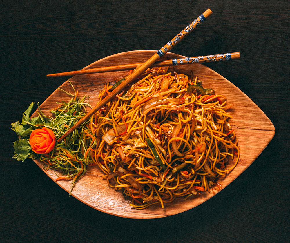 Noodles with vegetables and green garnish on side on wooden plate with chopsticks