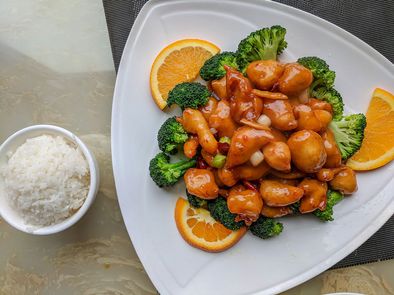 Bright bite-sized orange chicken on a layer of broccoli with orange slices garnish on a white plate and a side of rice